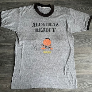 Może przedstawiać: Szary T-shirt z czarnymi rękawami typu ringer i nadrukiem "Alcatraz Reject" z przodu. Pod tekstem znajduje się postać z kreskówki z paskowaną chustką.