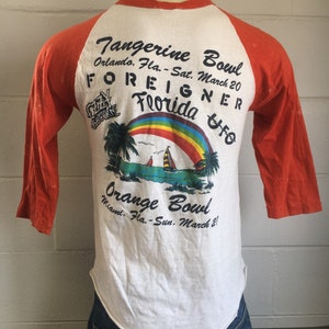 May include: A white t-shirt with orange sleeves featuring a graphic of a rainbow, palm trees, and a sailboat. The text "Tangerine Bowl" and "Orange Bowl" are printed on the shirt, along with the dates "Sat. March 20" and "Sun. March 21". The text "FOREIGNER" and "Florida" are also printed on the shirt.