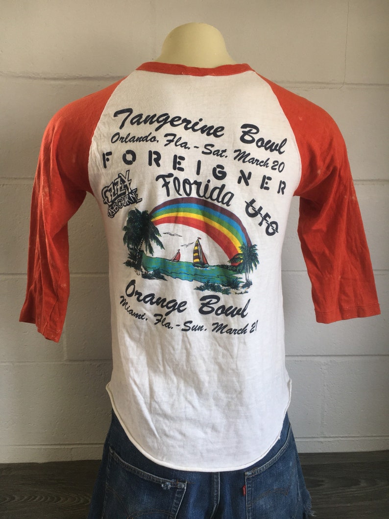 May include: A white t-shirt with orange sleeves featuring a graphic of a rainbow, palm trees, and a sailboat. The text "Tangerine Bowl" and "Orange Bowl" are printed on the shirt, along with the dates "Sat. March 20" and "Sun. March 21". The text "FOREIGNER" and "Florida" are also printed on the shirt.