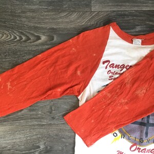 May include: A vintage baseball style t-shirt with orange sleeves and a white body. The front of the shirt features a graphic of a band with the text "Tangerine" and "Orange Bowl" printed on the shirt.