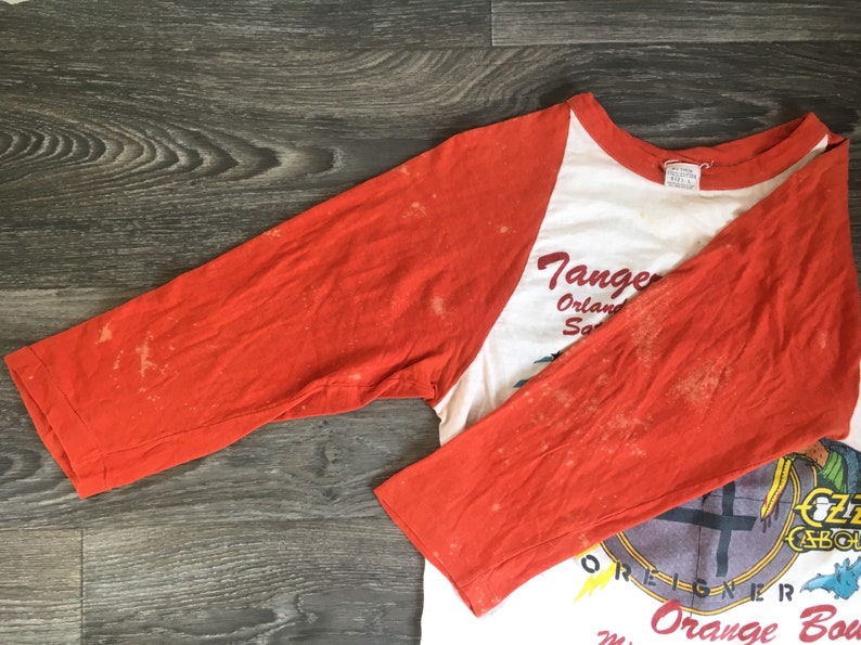 May include: A vintage baseball style t-shirt with orange sleeves and a white body. The front of the shirt features a graphic of a band with the text "Tangerine" and "Orange Bowl" printed on the shirt.
