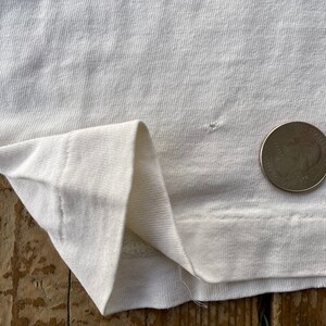 K&ouml;nnte beinhalten: Nahaufnahme eines wei&szlig;en T-Shirts mit einem kleinen Loch in der N&auml;he des unteren Saums. Eine US-amerikanische 25-Cent-M&uuml;nze liegt zum Gr&ouml;&szlig;envergleich auf dem Shirt.