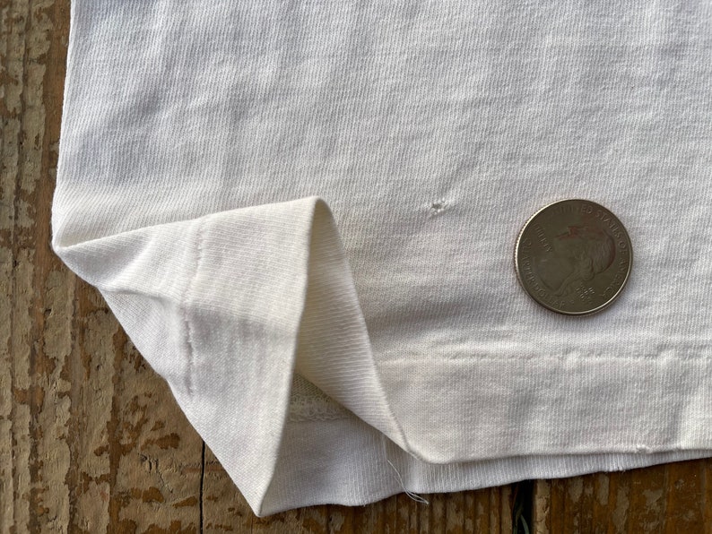 K&ouml;nnte beinhalten: Nahaufnahme eines wei&szlig;en T-Shirts mit einem kleinen Loch in der N&auml;he des unteren Saums. Eine US-amerikanische 25-Cent-M&uuml;nze liegt zum Gr&ouml;&szlig;envergleich auf dem Shirt.