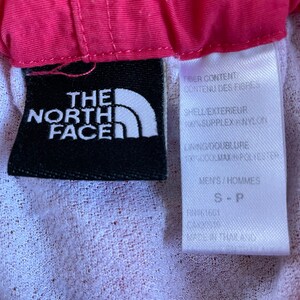 May include: A black and white label with the text "The North Face" and a white triangle logo. The label is attached to a pink and white garment. The label also includes the text "Fiber Content", "Contenu des Fibres", "Shell Exterieur", "100% Supplex Nylon", "Lining/Doublure", "100% Max Performance", "Men's/Hommes", "S-P", "FN61661", "THAILAND".