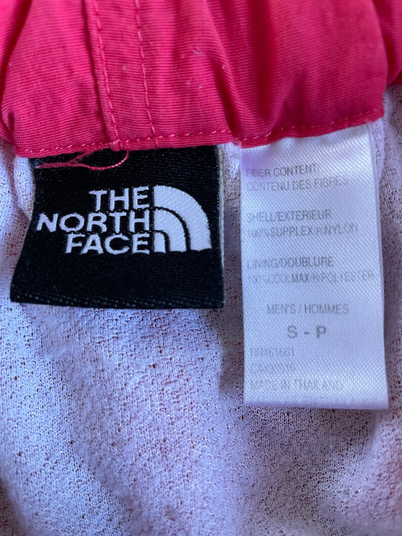 May include: A black and white label with the text "The North Face" and a white triangle logo. The label is attached to a pink and white garment. The label also includes the text "Fiber Content", "Contenu des Fibres", "Shell Exterieur", "100% Supplex Nylon", "Lining/Doublure", "100% Max Performance", "Men's/Hommes", "S-P", "FN61661", "THAILAND".