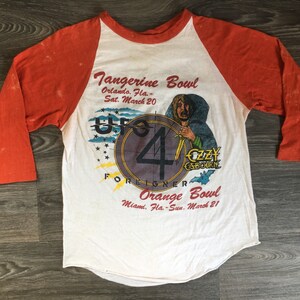 May include: A vintage white and orange baseball t-shirt with a graphic of a band called "UFO" and the text "Tangerine Bowl Orlando, Fla. - Sat. March 20" and "Orange Bowl Miami, Fla. - Sun. March 21".