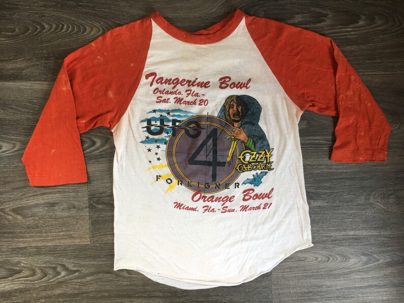 May include: A vintage white and orange baseball t-shirt with a graphic of a band called "UFO" and the text "Tangerine Bowl Orlando, Fla. - Sat. March 20" and "Orange Bowl Miami, Fla. - Sun. March 21".