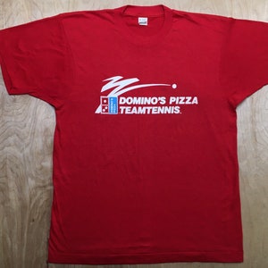 May include: Red t-shirt with a white graphic of a Domino's Pizza Team Tennis logo. The logo features a white tennis ball, a white tennis racket, and the text "Domino's Pizza Team Tennis."