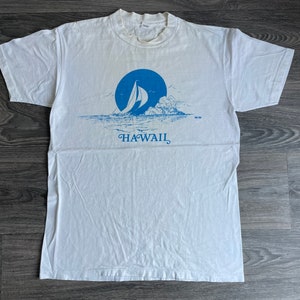 May include: White t-shirt with a blue graphic of a sailboat sailing under a blue moon with the word "HAWAII" printed below.
