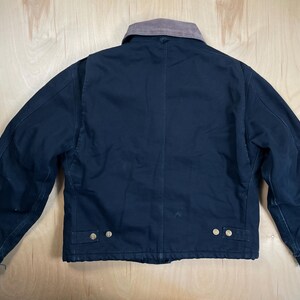 May include: A navy blue work jacket with a brown corduroy collar. The jacket has a button-up front, two chest pockets, and two lower pockets. The jacket is made of a durable material and is designed for work.