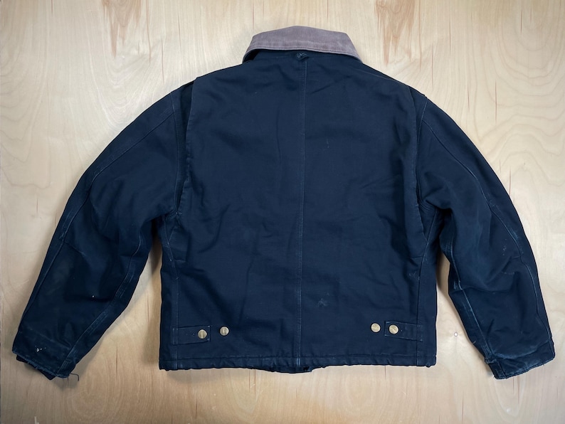May include: A navy blue work jacket with a brown corduroy collar. The jacket has a button-up front, two chest pockets, and two lower pockets. The jacket is made of a durable material and is designed for work.