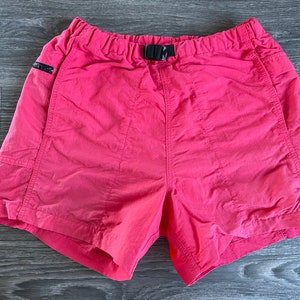 May include: Pink nylon shorts with a black elastic waistband and a black adjustable buckle. The shorts have two side pockets and two back pockets.