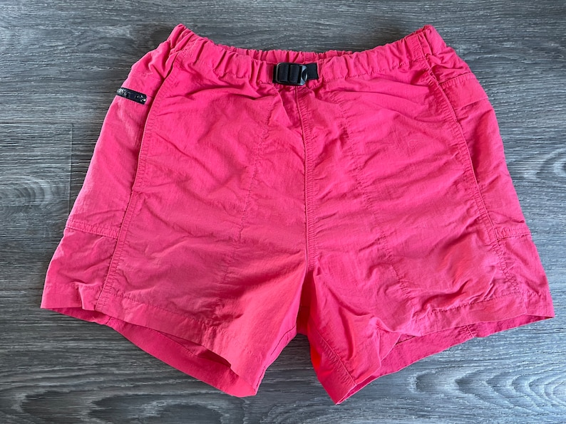 May include: Pink nylon shorts with a black elastic waistband and a black adjustable buckle. The shorts have two side pockets and two back pockets.