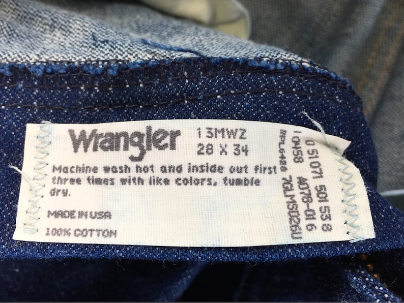 Puede incluir: Etiqueta de cuidado blanca con texto negro para jeans Wrangler. La etiqueta incluye la talla 28 x 34, el n&uacute;mero de estilo 13MWZ y las instrucciones de lavado. La etiqueta tambi&eacute;n indica "Hecho en EE. UU." y "100 % algod&oacute;n".