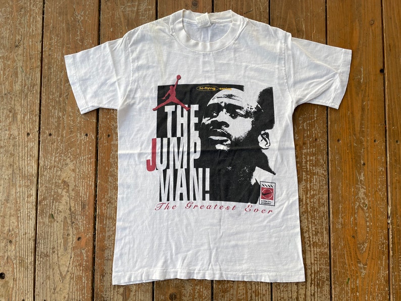 K&ouml;nnte beinhalten: Wei&szlig;es T-Shirt mit einer schwarz-wei&szlig;en Grafik von Michael Jordan, dem Text "The Jumpman" und den Worten "The Greatest Ever".