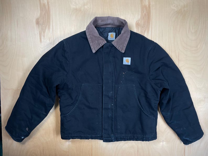 May include: Black Carhartt jacket with a brown corduroy collar and two front pockets. The jacket has the Carhartt logo on the chest and collar. The jacket is a workwear style.