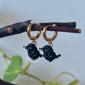 May include: Gold hoop earrings with dangling black beaded bird-shaped charms. The earrings are displayed on a dark brown twig against a soft, neutral background. The earrings have a delicate and artistic design.