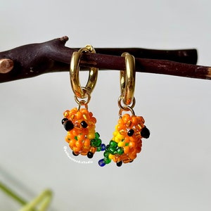 以下が含まれることがあります： カラフルなビーズでできた鳥のチャームが付いた、ゴールドのフープイヤリングのペア。鳥はオレンジ、イエロー、グリーン、ブルーのビーズで作られています。