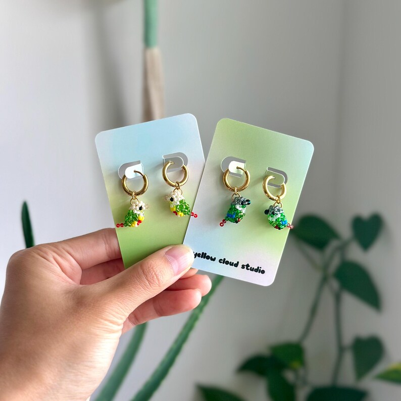 May include: Two pairs of beaded bird earrings on white and green card stock with the text "yellow cloud studio". The earrings are gold hoops with a small beaded bird charm hanging from each hoop. One pair of earrings features a green and white bird, and the other pair features a blue and green bird.