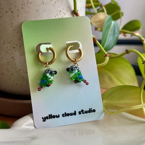May include: A pair of gold hoop earrings with green, blue, black, and white beaded bird charms hanging from them. The earrings are displayed on a card with the text "yellow cloud studio".