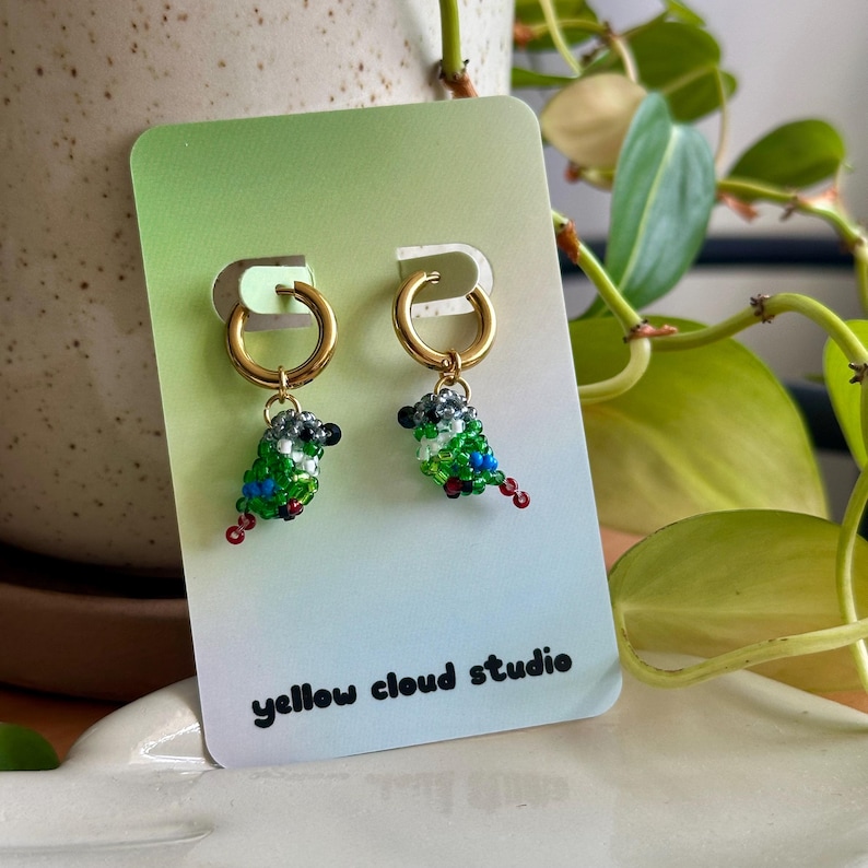 May include: A pair of gold hoop earrings with green, blue, black, and white beaded bird charms hanging from them. The earrings are displayed on a card with the text "yellow cloud studio".