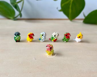 May include: A collection of seven small, beaded bird figurines. The birds are in various colors, including green, yellow, red, white, and pink. The birds are arranged in a line on a light brown surface.
