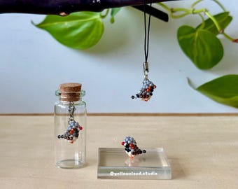 May include: A clear glass bottle with a cork stopper, a beaded bird charm inside, and a beaded bird charm hanging from a black cord. A third beaded bird sits on a clear acrylic block. The birds are gray, orange, black, and white.