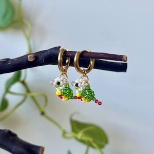 May include: A pair of gold hoop earrings with small beaded bird charms. The birds are green, white, red, and yellow.