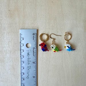 May include: A pair of beaded bird earrings. One earring features a red, blue, and yellow bird, while the other earring features a green, yellow, and white bird. The earrings are approximately 1 inch long.