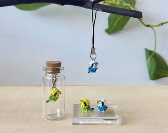 May include: A clear glass bottle with a cork stopper and a beaded bird charm. Two beaded bird figurines, one yellow and green, the other blue and white, sit on a clear stand. A beaded bird charm hangs from a black cord. The text "@yellowcloud.studio" is visible.
