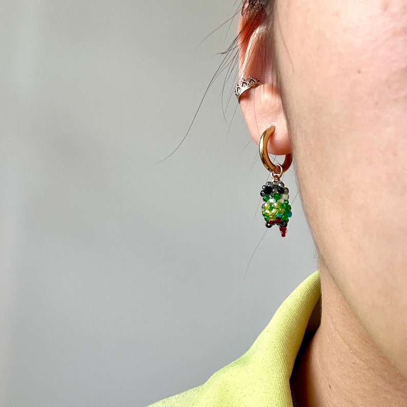 May include: A gold hoop earring with a beaded parrot charm. The parrot is green, black, white, and red.