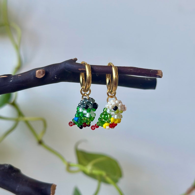 May include: A pair of gold hoop earrings with small beaded bird charms. One bird is green, white, and red, and the other is green, white, and yellow.