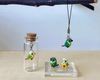 May include: Three small beaded parakeet figurines. One parakeet is hanging from a black string, and two are on a small white display stand. The parakeets are green, white, and yellow.