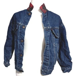 May include: A blue denim jacket featuring a red and black plaid collar. The jacket has a button-front closure with metallic buttons, long sleeves, and two chest pockets with buttoned flaps. The denim fabric has a medium wash finish throughout.