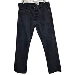 May include: Dark gray denim jeans with a button fly, five-pocket design, and a small orange accent. The jeans are hanging on a metal hanger against a white background. The DC logo is visible on the button.