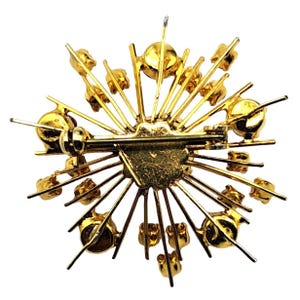 Puede incluir: Broche dorado con dise&ntilde;o de estrella. El broche presenta un pasador y cierre central, con l&iacute;neas radiales y peque&ntilde;os acentos redondos de color dorado. La parte posterior del broche es visible, mostrando el mecanismo del pasador.