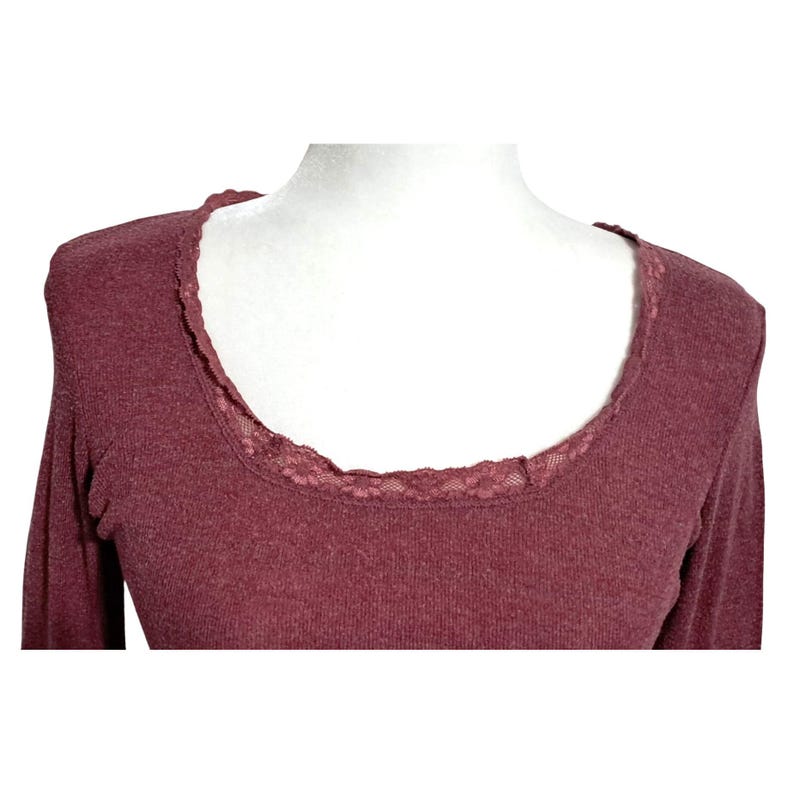 May include: A long-sleeved, burgundy-colored shirt with a scoop neckline. The neckline is trimmed with delicate lace. The shirt is displayed on a white mannequin, showcasing the garment's shape and design. The fabric appears to be a soft knit.