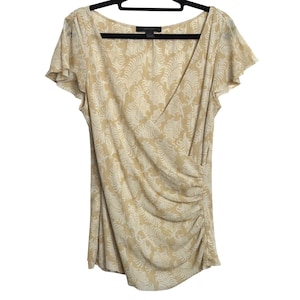 May include: A beige short-sleeve top with a wrap-style front and a subtle, all-over floral pattern. The top has a V-neckline and is hanging on a black hanger. The fabric appears soft and lightweight, suitable for warmer weather.