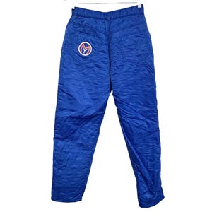 May include: Bright blue quilted snow pants with a high waist and a straight leg. The pants feature two back pockets and a circular patch with a stylized "M" logo. The pants are made of a shiny, water-resistant material.