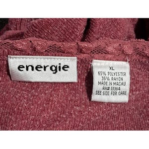 May include: Close-up of a burgundy-colored top with the brand label "energie" visible. The top has a textured knit fabric and a lace trim. A tag indicates the size is XL and the fabric content is 65% polyester and 35% rayon.