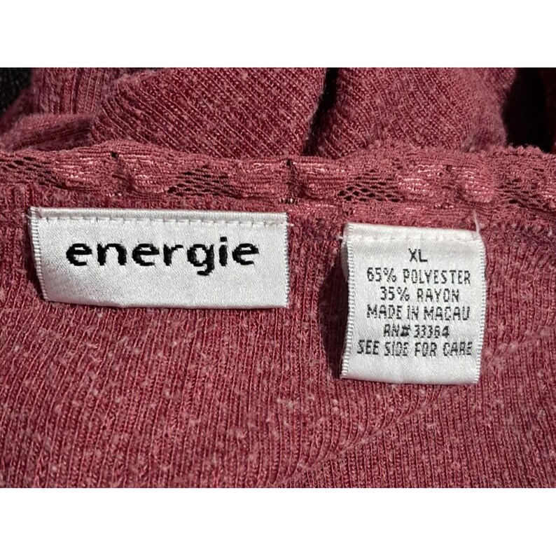 May include: Close-up of a burgundy-colored top with the brand label "energie" visible. The top has a textured knit fabric and a lace trim. A tag indicates the size is XL and the fabric content is 65% polyester and 35% rayon.