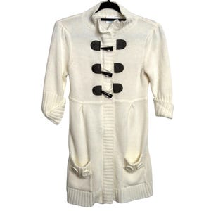 May include: Cream-colored long cardigan sweater with three-quarter sleeves and a button-up closure. The sweater features three dark brown toggle closures and two front pockets. The knitwear has a ribbed texture along the center and cuffs.