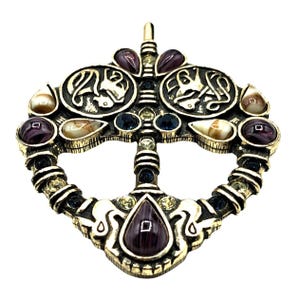 May include: A gold-toned pendant with a Celtic knot design and various colored gemstones. The pendant features a central design with two smaller circles on either side, each with a stylized animal figure. The pendant is adorned with purple, blue, and white gemstones.