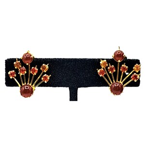 Puede incluir: Un par de pendientes dorados con un dise&ntilde;o en forma de abanico. Cada pendiente presenta una piedra grande, redonda, de color marr&oacute;n rojizo en la base, con piedras rojas y naranjas m&aacute;s peque&ntilde;as que se extienden en abanico. Los pendientes se exhiben sobre una superficie negra.