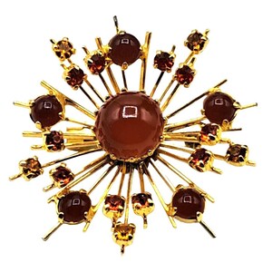 Puede incluir: Broche dorado con dise&ntilde;o de rayos de sol. El centro presenta una piedra grande, redonda, de color marr&oacute;n rojizo, rodeada de l&iacute;neas doradas radiantes. Piedras m&aacute;s peque&ntilde;as de color marr&oacute;n rojizo y &aacute;mbar se intercalan a lo largo de los rayos, creando un efecto brillante. Una joya cl&aacute;sica.
