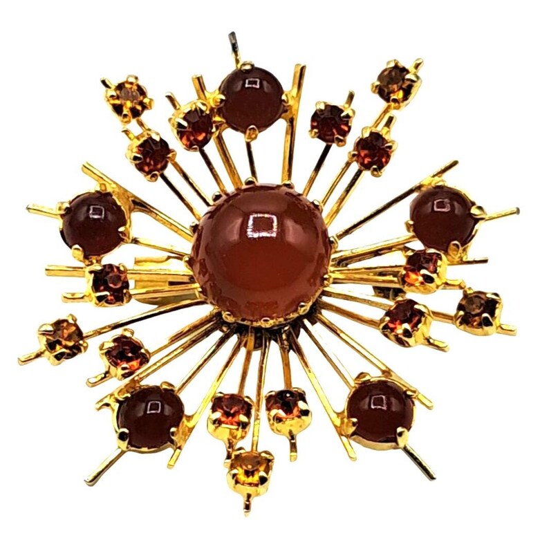 Puede incluir: Broche dorado con dise&ntilde;o de rayos de sol. El centro presenta una piedra grande, redonda, de color marr&oacute;n rojizo, rodeada de l&iacute;neas doradas radiantes. Piedras m&aacute;s peque&ntilde;as de color marr&oacute;n rojizo y &aacute;mbar se intercalan a lo largo de los rayos, creando un efecto brillante. Una joya cl&aacute;sica.