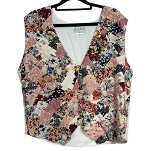 May include: A patchwork vest with a V-neck design. The vest features a variety of floral patterns in shades of pink, cream, burgundy, and navy blue. It has a gold-colored clasp closure and a white lining. The vest is hanging on a black hanger.