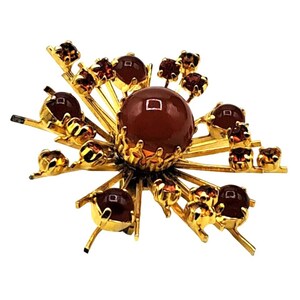 Puede incluir: Broche dorado con dise&ntilde;o de estrella. El centro presenta una piedra grande, redonda, de color rojo oscuro, rodeada de piedras m&aacute;s peque&ntilde;as y detalles dorados. La broche tiene una est&eacute;tica vintage.