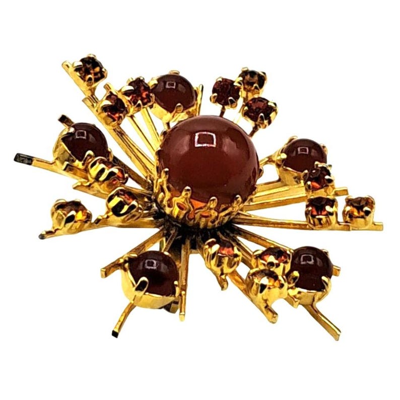 Puede incluir: Broche dorado con dise&ntilde;o de estrella. El centro presenta una piedra grande, redonda, de color rojo oscuro, rodeada de piedras m&aacute;s peque&ntilde;as y detalles dorados. La broche tiene una est&eacute;tica vintage.