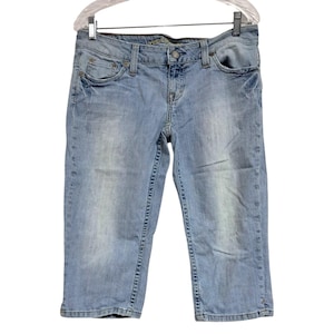 May include: Light blue denim capri pants with a classic five-pocket design. The capris have a button and zipper closure, belt loops, and a faded wash. The pants are hanging on a clear plastic hanger.